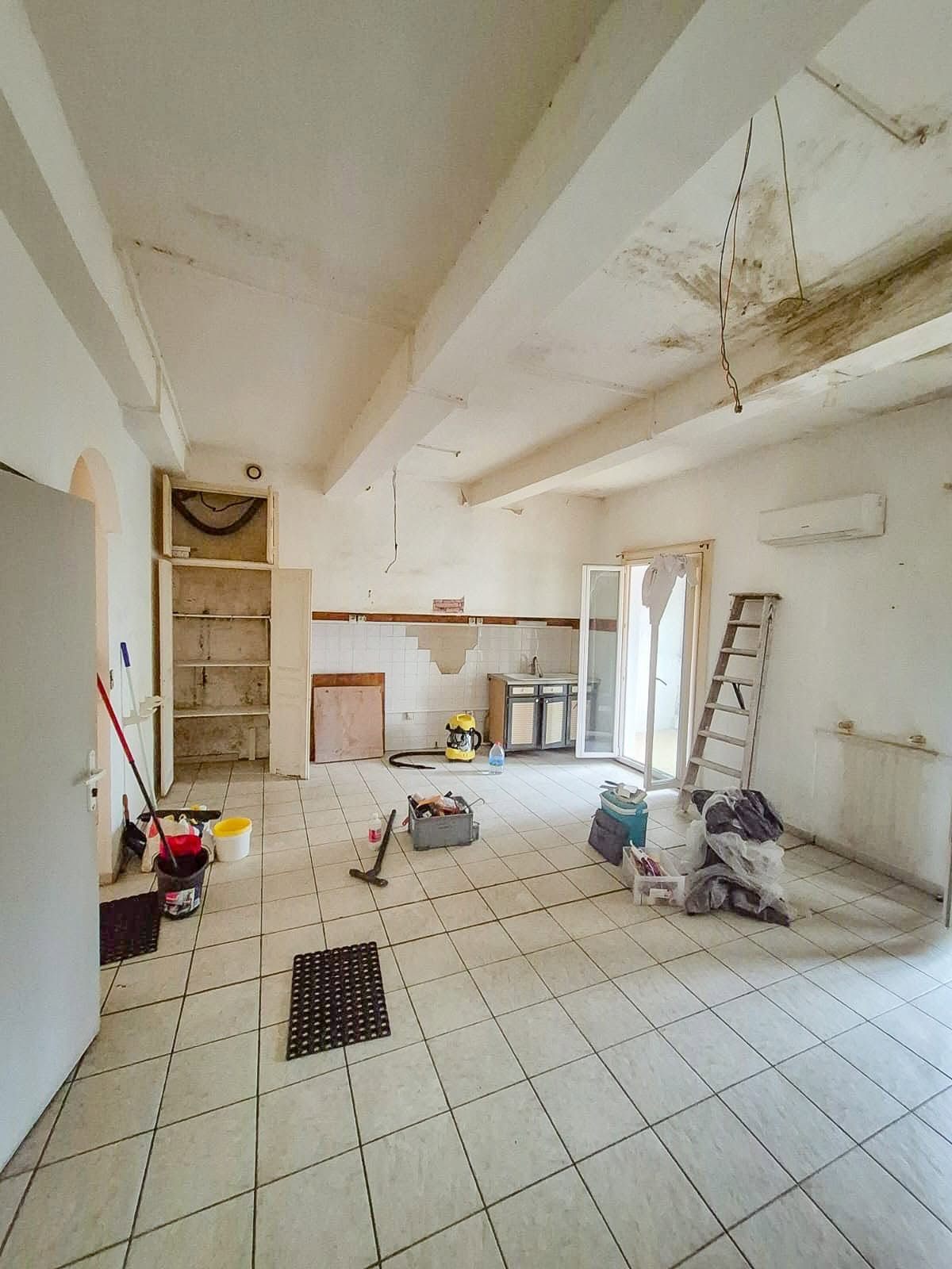 Grand séjour à poutres apparentes avant travaux : murs et plafond marqués, matériel de chantier au sol
