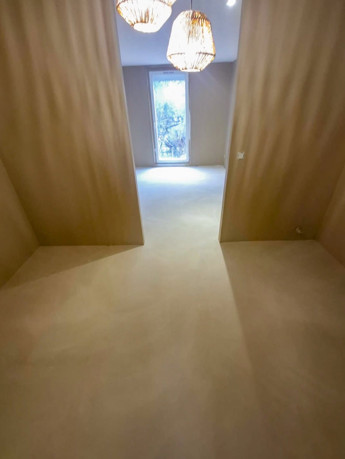Couloir et pièce traités en enduit décoratif beige continu sur les murs et le sol, éclairés par des suspensions en rotin