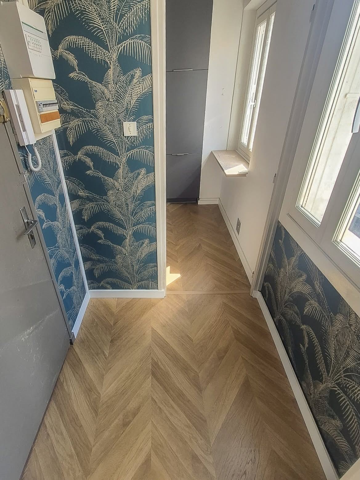 Entrée tapissée d'un papier peint à motif de palmes crème sur fond bleu canard, posée sur un parquet à chevrons