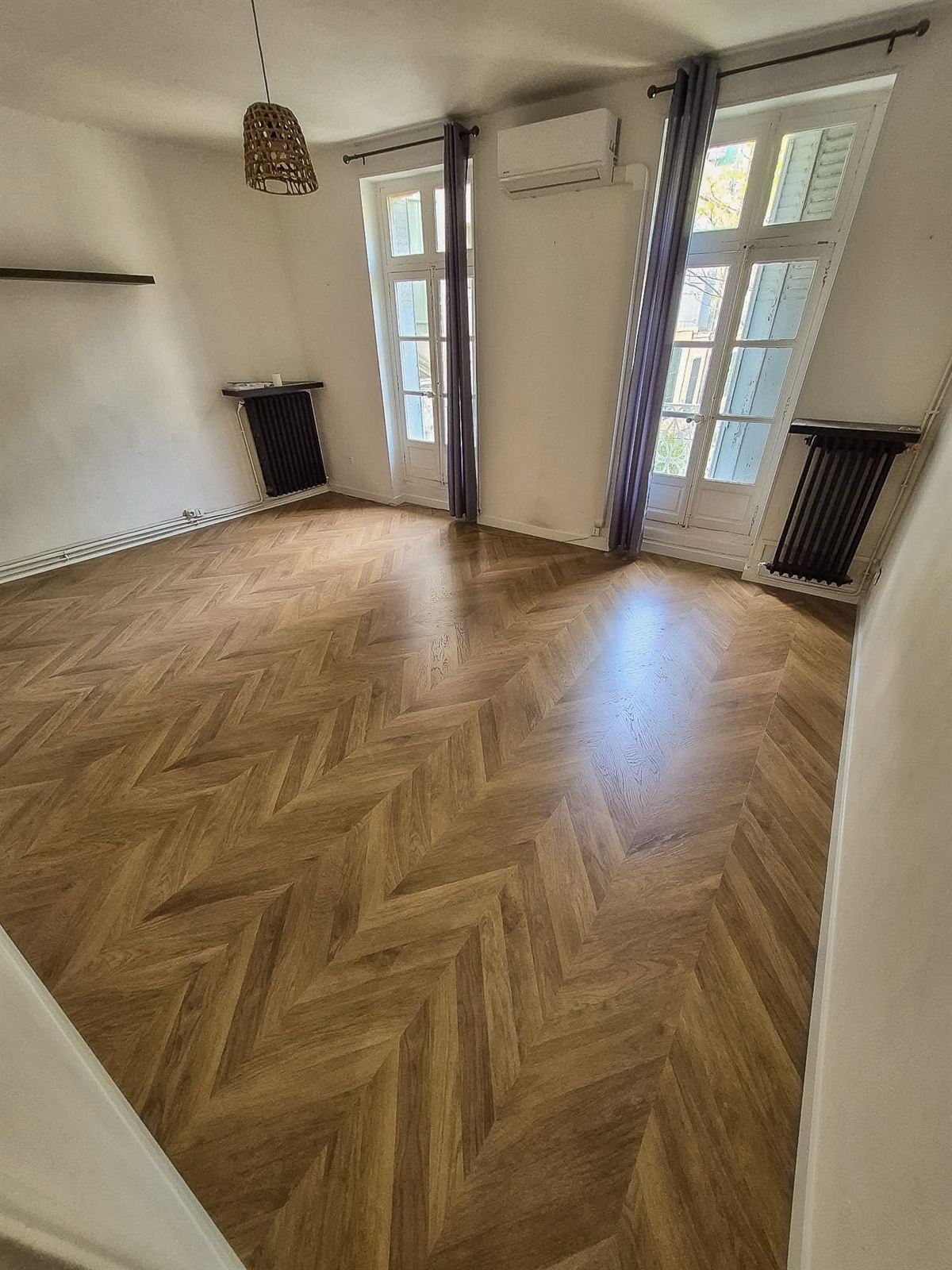 Pièce vide au parquet à chevrons, avec deux portes-fenêtres à petits carreaux et radiateurs en fonte
